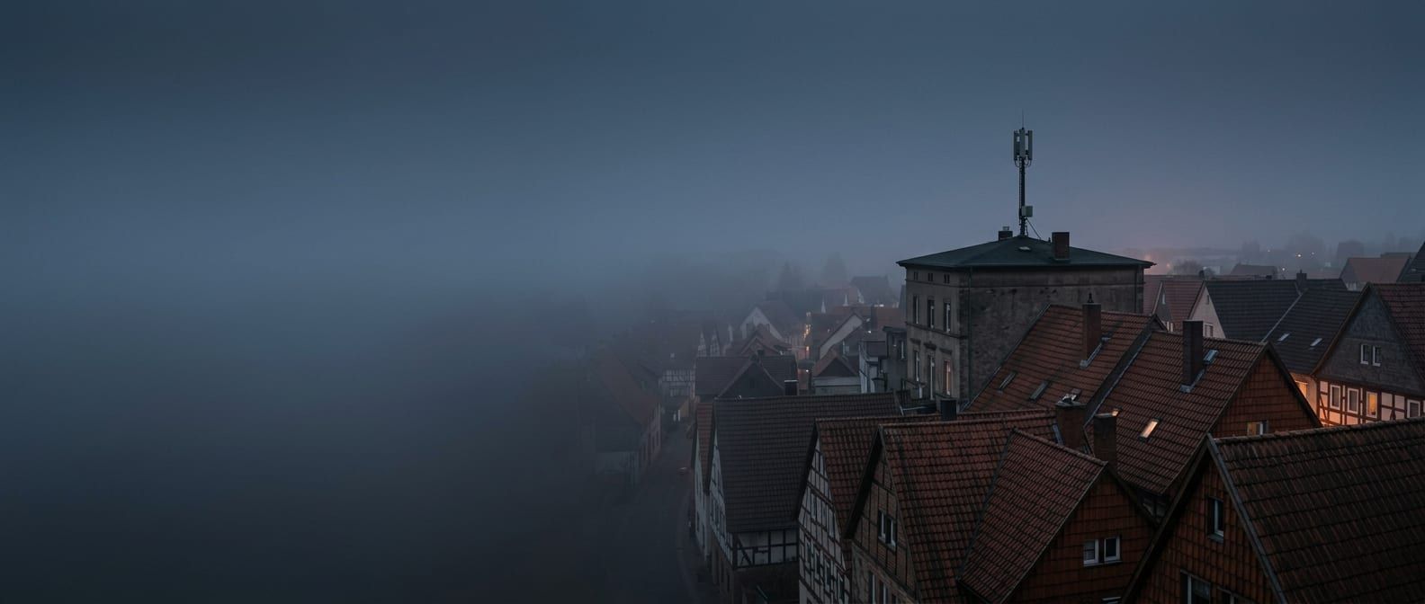Ziegeldächer einer Kleinstadt im Abendnebel, dahinter ein höheres Wohngebäude mit Mobilfunkanlage auf dem Flachdach