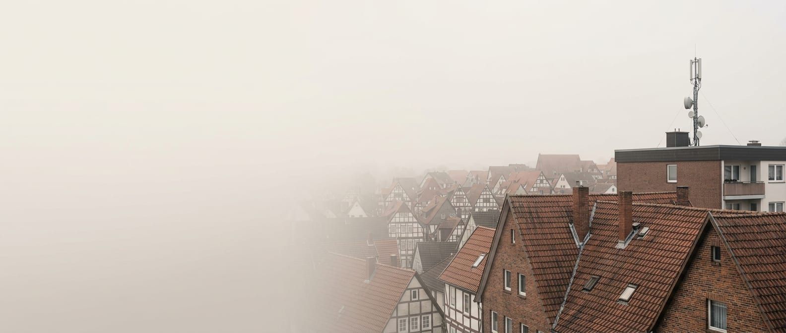 Ziegeldächer einer Kleinstadt im Abendnebel, dahinter ein höheres Wohngebäude mit Mobilfunkanlage auf dem Flachdach